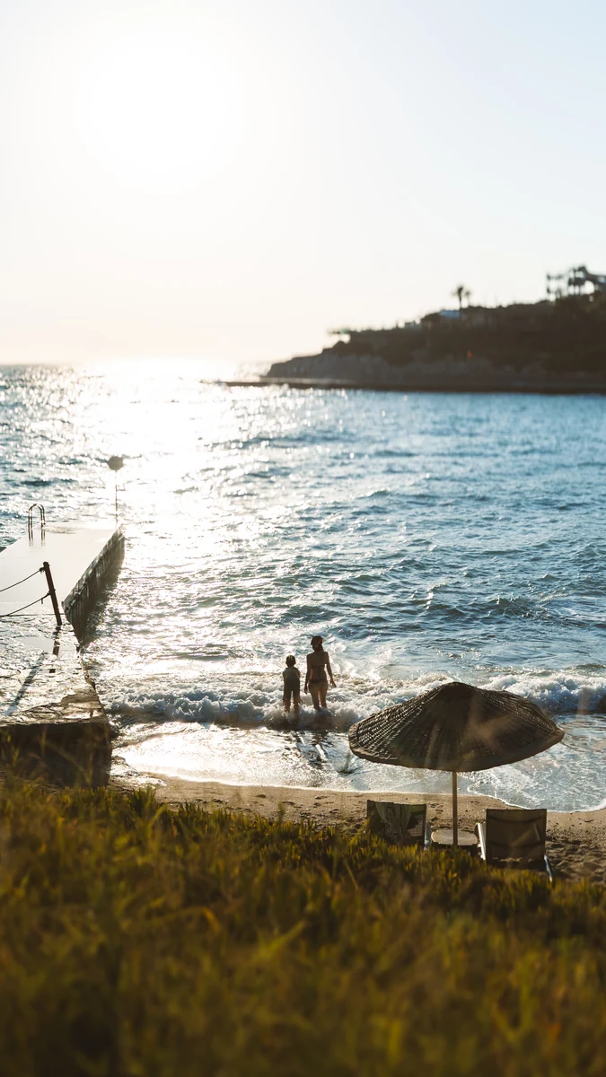 Two figures at the waterline beside a sunlit pier